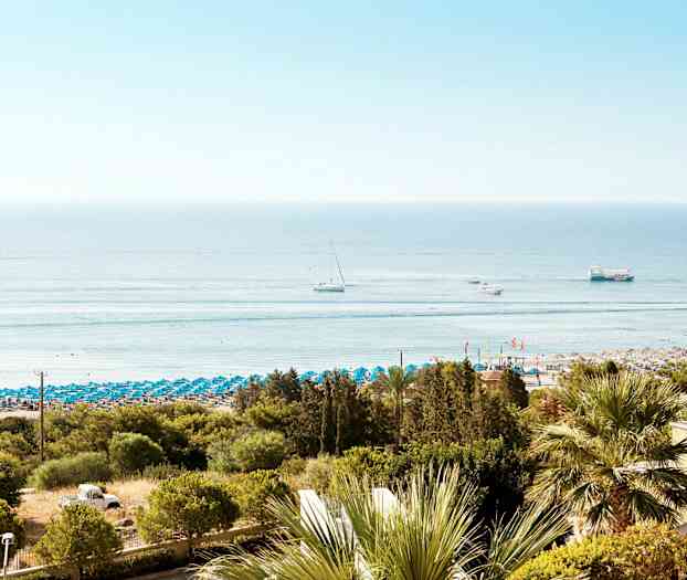 En strand med blå parasoller og havet i baggrunden. Flere både ses på vandet. (AI-genereret tekst)