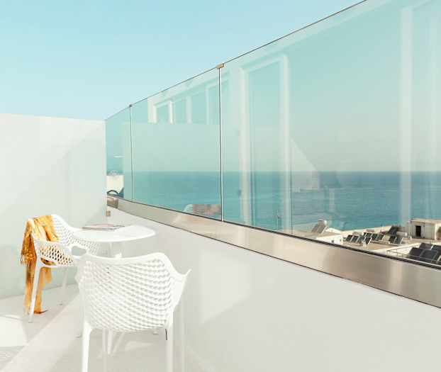 En hvid balkon med to stole og et bord, med udsigt over havet og bygninger. (AI-genereret tekst)