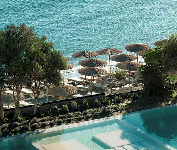 Udsigt over poolen, træer og stranden med parasoller og liggestole mod havet. (AI-genereret tekst)