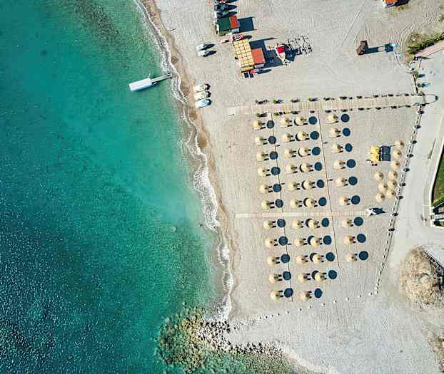 Luftfoto af en strand med parasoller og liggestole ved siden af klart blåt vand. (AI-genereret tekst)