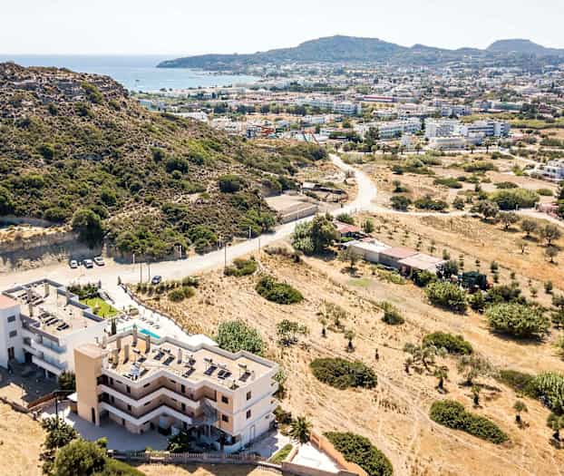 Luftfoto af en bygning nær en bjergskråning med udsigt over en by og havet. (AI-genereret tekst)