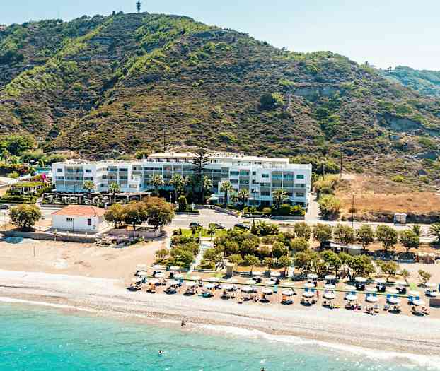 Strand med liggestole og parasoller, bagved ligger en bygning ved en bjergskråning. (AI-genereret tekst)