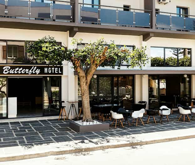 Forsiden af et hotel med skiltet "Butterfly Hotel", en terrasse med borde og stole samt et træ foran bygningen. (AI-genereret tekst)