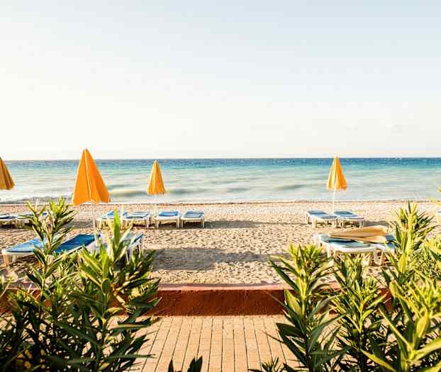 En strand med liggestole og gule parasoller ved et blåt hav, omgivet af planter. (AI-genereret tekst)