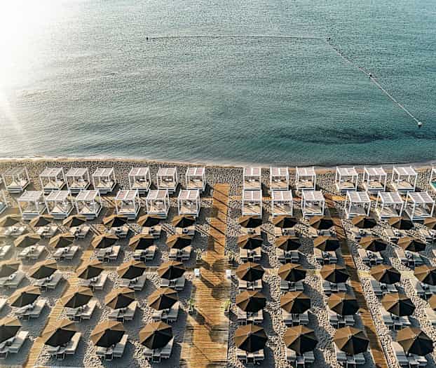 Et blik over en strand med rækker af parasoller, liggestole og hvide solskærme tæt på havet. (AI-genereret tekst)