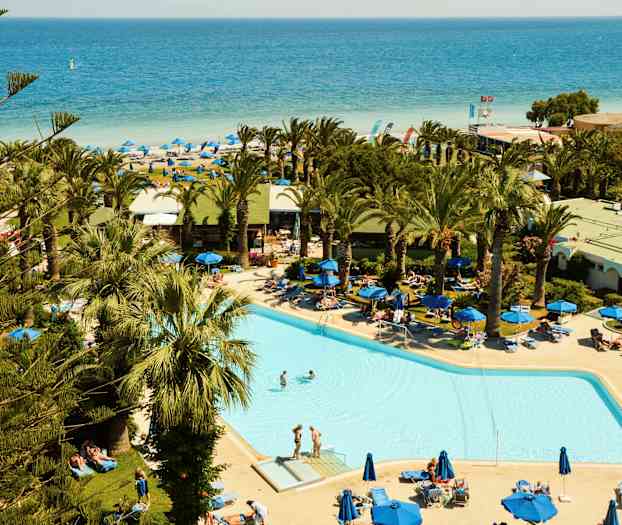 En pool omgivet af liggestole og palmer, med en sandstrand og havet i baggrunden. (AI-genereret tekst)