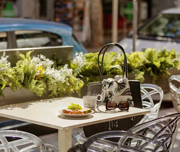 Bord med croissant, kop og solbriller på udendørs café med blomsterdekorationer. (AI-genereret tekst)