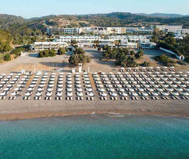 Liggestole og parasoller i flere rækker på en sandstrand med turkis vand, omgivet af grønne områder og bygninger i baggrunden. (AI-genereret tekst)