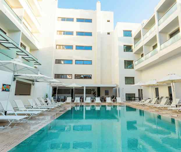 Billede af et hotelområde med en swimmingpool omgivet af liggestole og parasoller ovenover. Baggrunden viser hvide bygninger med altaner. (AI-genereret tekst)