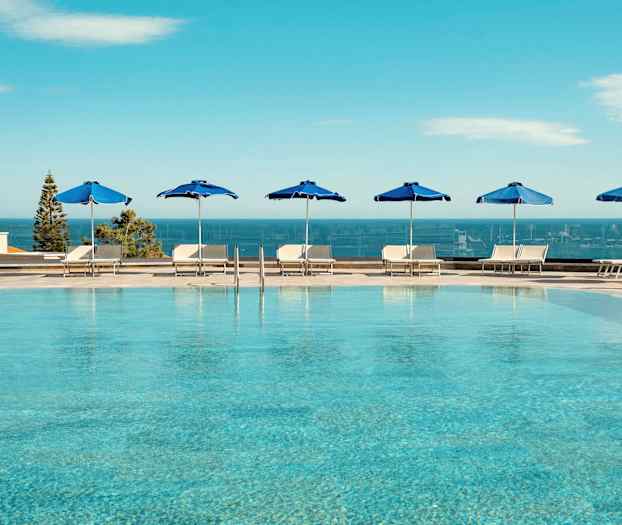 Udendørs pool med blå parasoller og udsigt over havet. (AI-genereret tekst)
