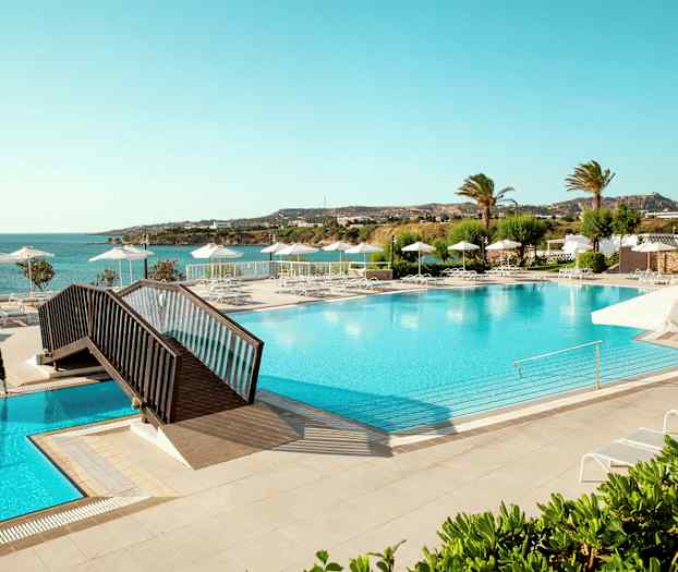 Stor pool med liggestole og parasoller, udsigt mod havet og grønne områder. (AI-genereret tekst)