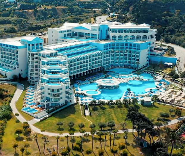 Stor hotelbygning med flere blå altaner og pools, omgivet af grønt. (AI-genereret tekst)