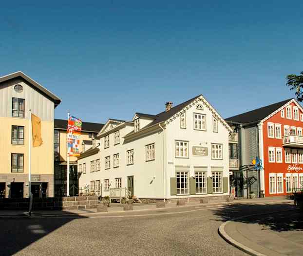 Billede af farverige bygninger med hotel og restaurant nær en gade med fortov. (AI-genereret tekst)