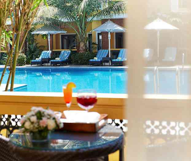 Billedet viser en pool med solsenge og parasoller samt et nærbillede af et bord med drikkevarer og blomster i forgrunden. (AI-genereret tekst)