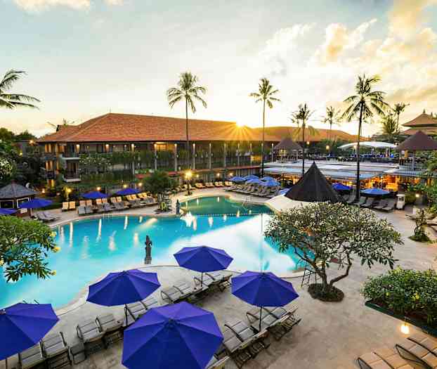 Billede af en resort med en pool, omgivet af liggestole og parasoller samt grønne palmer i baggrunden. (AI-genereret tekst)