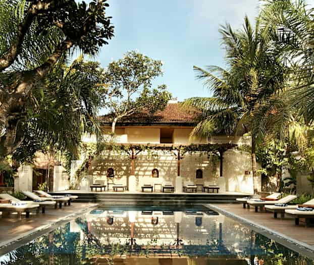 En udendørs pool omgivet af liggestole og træer med en bygning i baggrunden. (AI-genereret tekst)