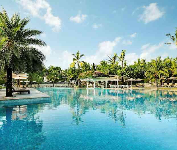 En stor pool omgivet af liggestole, parasoller og palmer, med en klar blå himmel i baggrunden. (AI-genereret tekst)