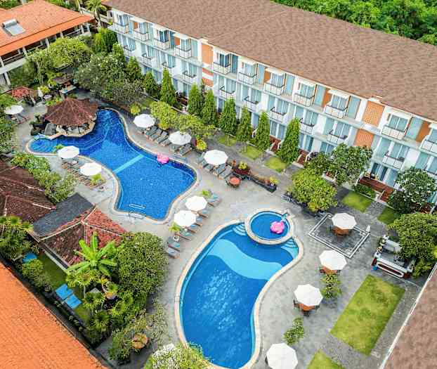 Et fugleperspektiv over et hotelområde med to swimmingpools, liggestole og parasoller, omgivet af beplantning og bygninger. (AI-genereret tekst)