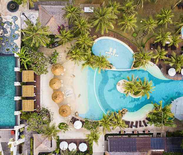 Luftfoto af en poolfacilitet med liggestole, parasoller og frodige palmer. (AI-genereret tekst)