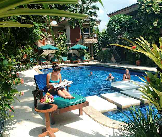 Et poolområde med liggestole, parasoller og frodige planter. En person sidder i en liggestol ved et bord med frugt, mens andre svømmer i poolen. (AI-genereret tekst)