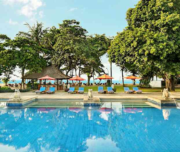 En udendørs pool omgivet af liggestole, parasoller og træer med havet i baggrunden. (AI-genereret tekst)