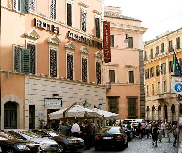 En gadescene med en hotelbygning med beige facade og mørke skodder. Foran bygningen står flere parkede biler og en café med parasoller. (AI-genereret tekst)