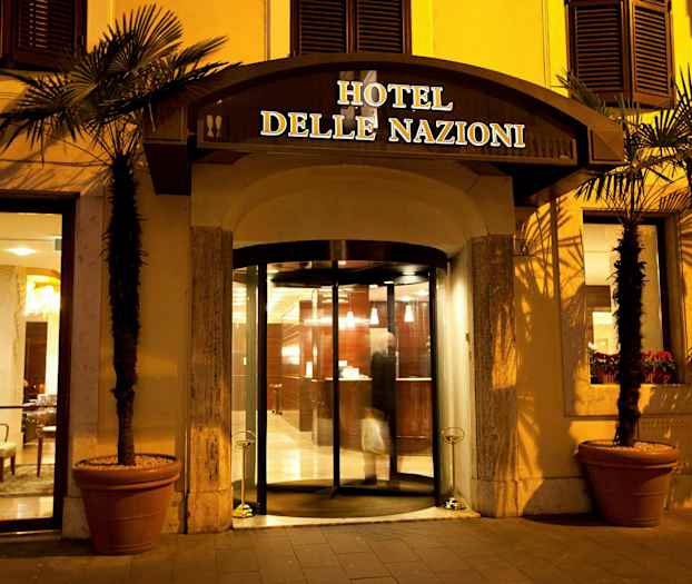 Indgangen til Hotel delle Nazioni med glasdøre og belysning om aftenen. (AI-genereret tekst)