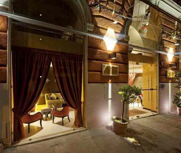 Billede af indgangen til en bygning med oplyst facade, store glasvinduer med røde gardiner og potteplanter. (AI-genereret tekst)