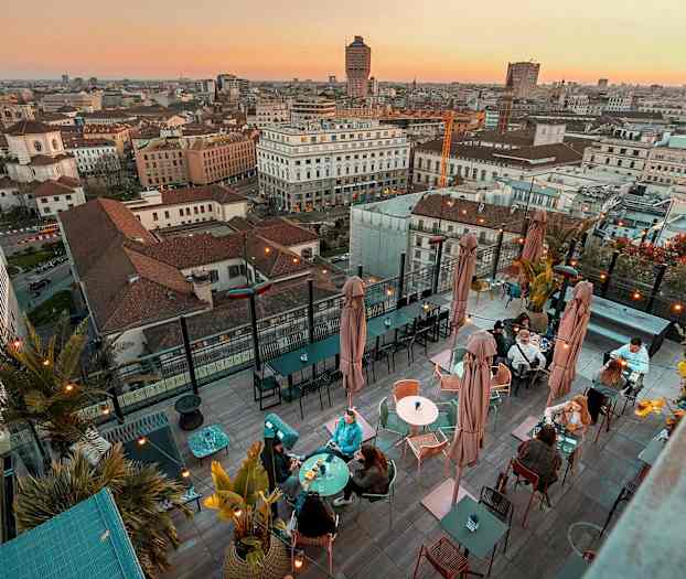 En tagterrasse med borde og siddepladser, omgivet af en panoramaudsigt over en by ved solnedgang. (AI-genereret tekst)