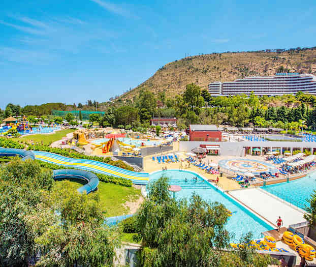 Billedet viser en stor udendørs vandpark med pools, vandrutsjebaner og omgivende solsenge. En stor bygning og skrånende terræn ses i baggrunden. (AI-genereret tekst)