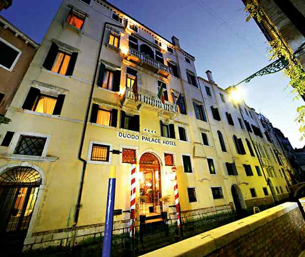 Lejlighedsbygning i tre etager med skilt Duodo Palace Hotel, omgivet af en kanal og en gangsti. (AI-genereret tekst)