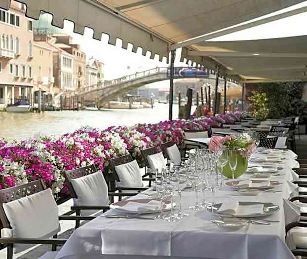 Et dækket middagbord med hvide duge placeret under en markise ved en kanal fyldt med både og dekoreret med farverige blomster langs kanten. (AI-genereret tekst)