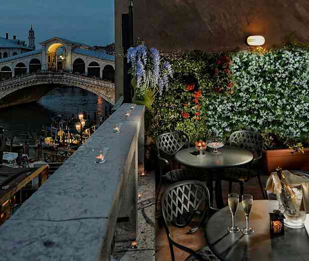 Billedet viser en terrasse med borde og stole, udsigt over Rialtobroen i Venedig, blomster og lys dekorerer området. (AI-genereret tekst)