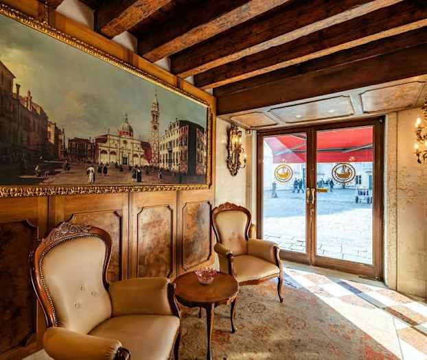 Indendørs scene med to stole og et bord foran en væg med et maleri, med udsigt til en ydre gennem glasdøre. (AI-genereret tekst)