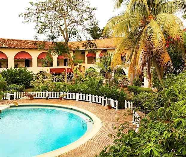 En rund pool omgivet af et stenbelagt område og hvide hegn. I baggrunden er en bygning med buede vinduer og røde markiser samt grønne omgivelser. (AI-genereret tekst)