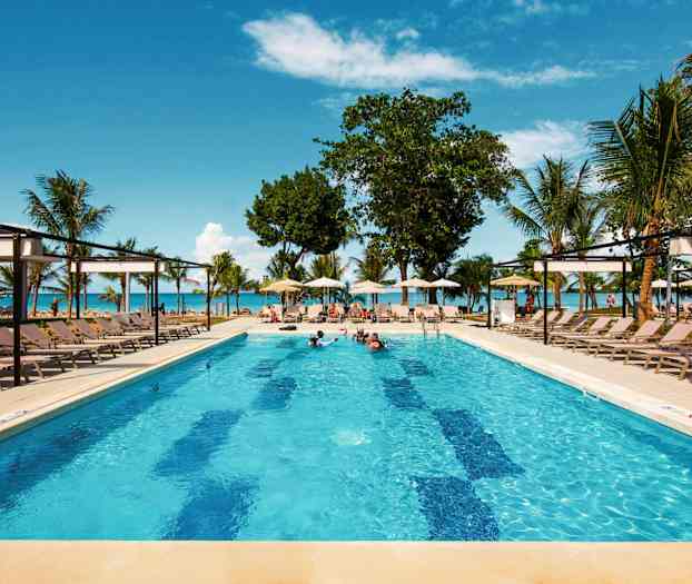 En pool med liggestole omkring, omgivet af palmer og med udsigt til havet og blå himmel. (AI-genereret tekst)