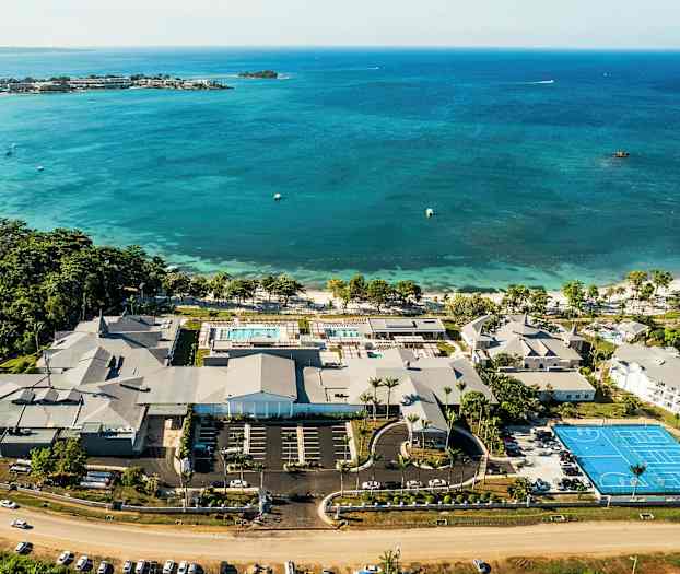 Luftfoto af en kystresort med pools, bygninger og en tennisbane ved havet. (AI-genereret tekst)