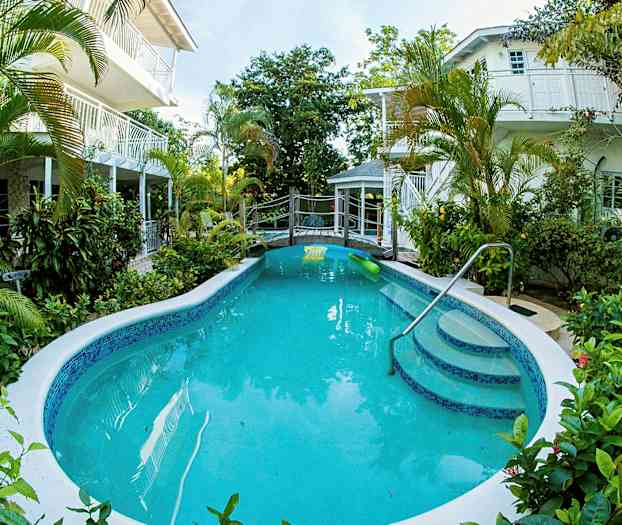 En udendørs pool omgivet af grønne planter med bygninger i baggrunden. (AI-genereret tekst)