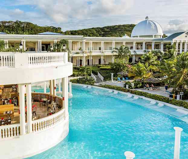 En stor bygning med hvide søjler og kuppel ved en pool omgivet af palmer og liggestole. (AI-genereret tekst)