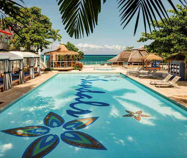 En pool med omkringliggende liggestole og en bygning med stråtag synlig ved havet. (AI-genereret tekst)