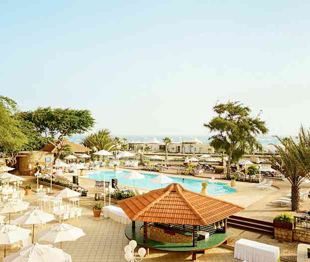 Billede af et udendørs område ved en pool med parasoller og træer under klar himmel. (AI-genereret tekst)