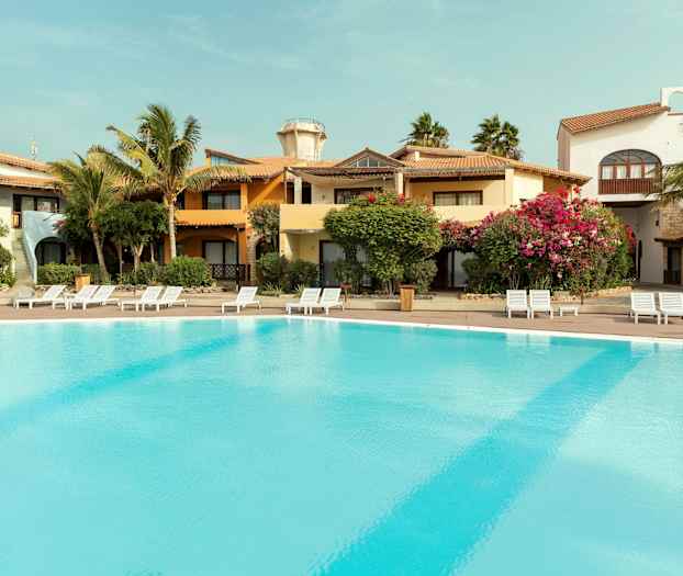 Udendørs pool ved ferieanlæg med omkringliggende bygninger og beplantning. (AI-genereret tekst)