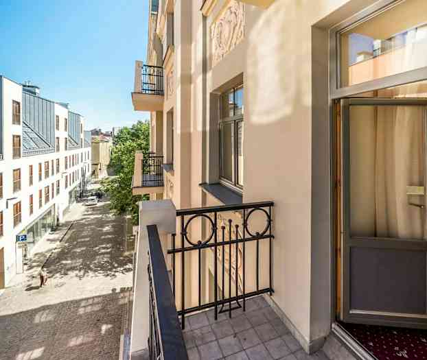 Billedet viser en altan på en lys bygning med udsigt over en brolagt gade og en moderne bygning med store vinduer på den anden side af gaden. (AI-genereret tekst)