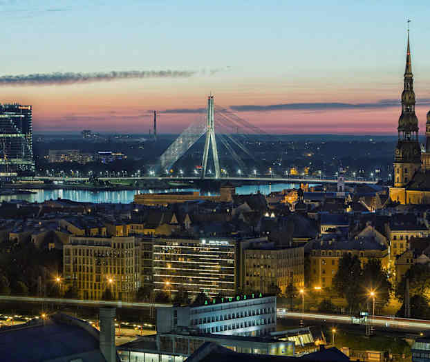Panoramabillede af en by i skumringen, med en flod, oplyste bygninger og en bro i baggrunden. (AI-genereret tekst)