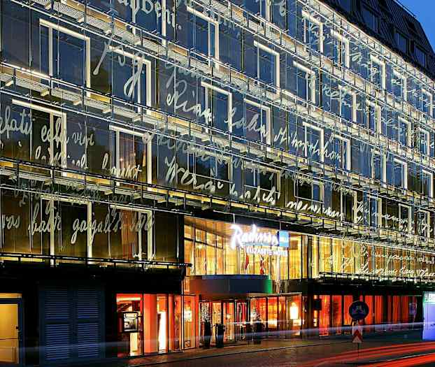 En moderne bygning med glasfacade oplyst om aftenen, dekoreret med skrifttyper. Indgangen til en hotelkæde er tydeligt synlig. (AI-genereret tekst)