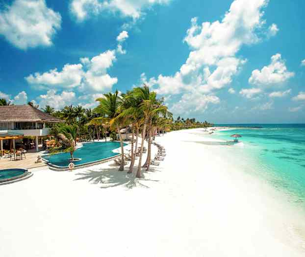 Billede af en hvid sandstrand med turkis vand, liggestole, parasoller og palmer ved et hotelområde. (AI-genereret tekst)