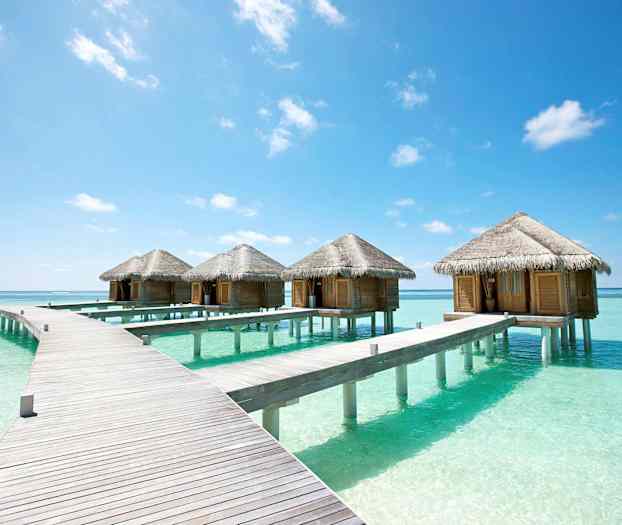 En række bungalows på pæle i det klare turkise vand under en blå himmel. (AI-genereret tekst)