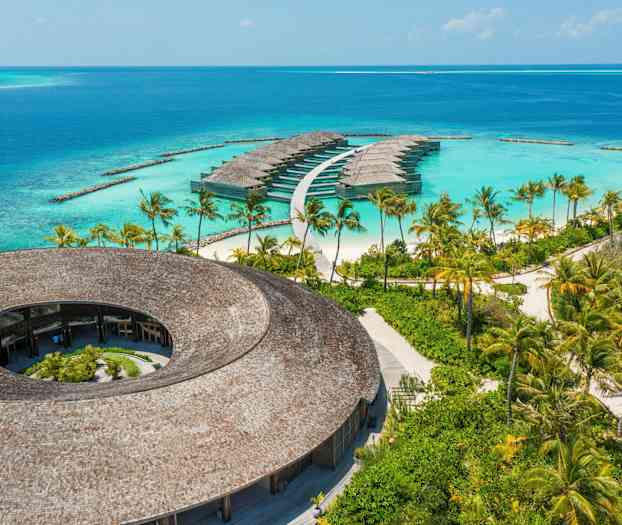 Udsigt over en rund bygning med trætak og vandvillaer omgivet af turkisblåt hav. (AI-genereret tekst)