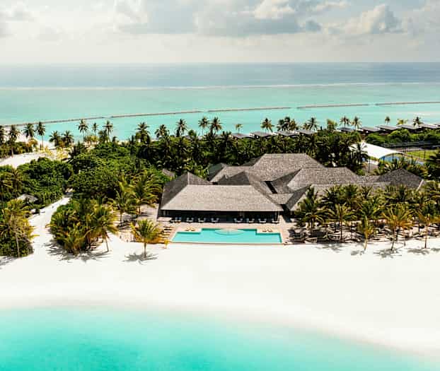 Luftfoto af en sandstrand med en pool, omgivet af palmer og bygninger ved havet. (AI-genereret tekst)