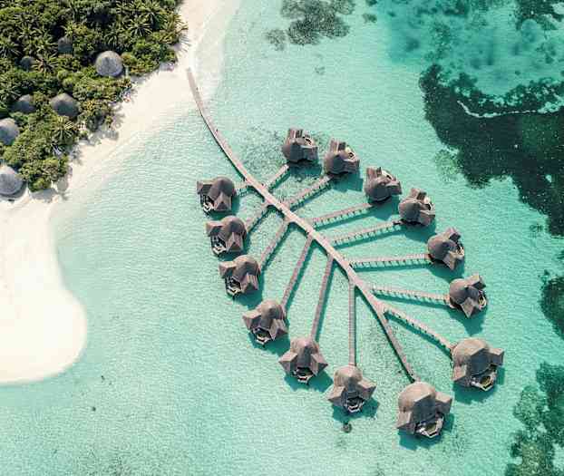 Luftfoto af bungalower på pæle over klart blåt vand med en nærliggende sandstrand og frodig vegetation. (AI-genereret tekst)